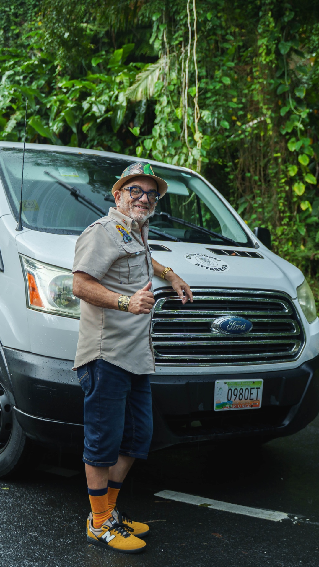 Person in casual outfit stands in front of a van on a tropical road.