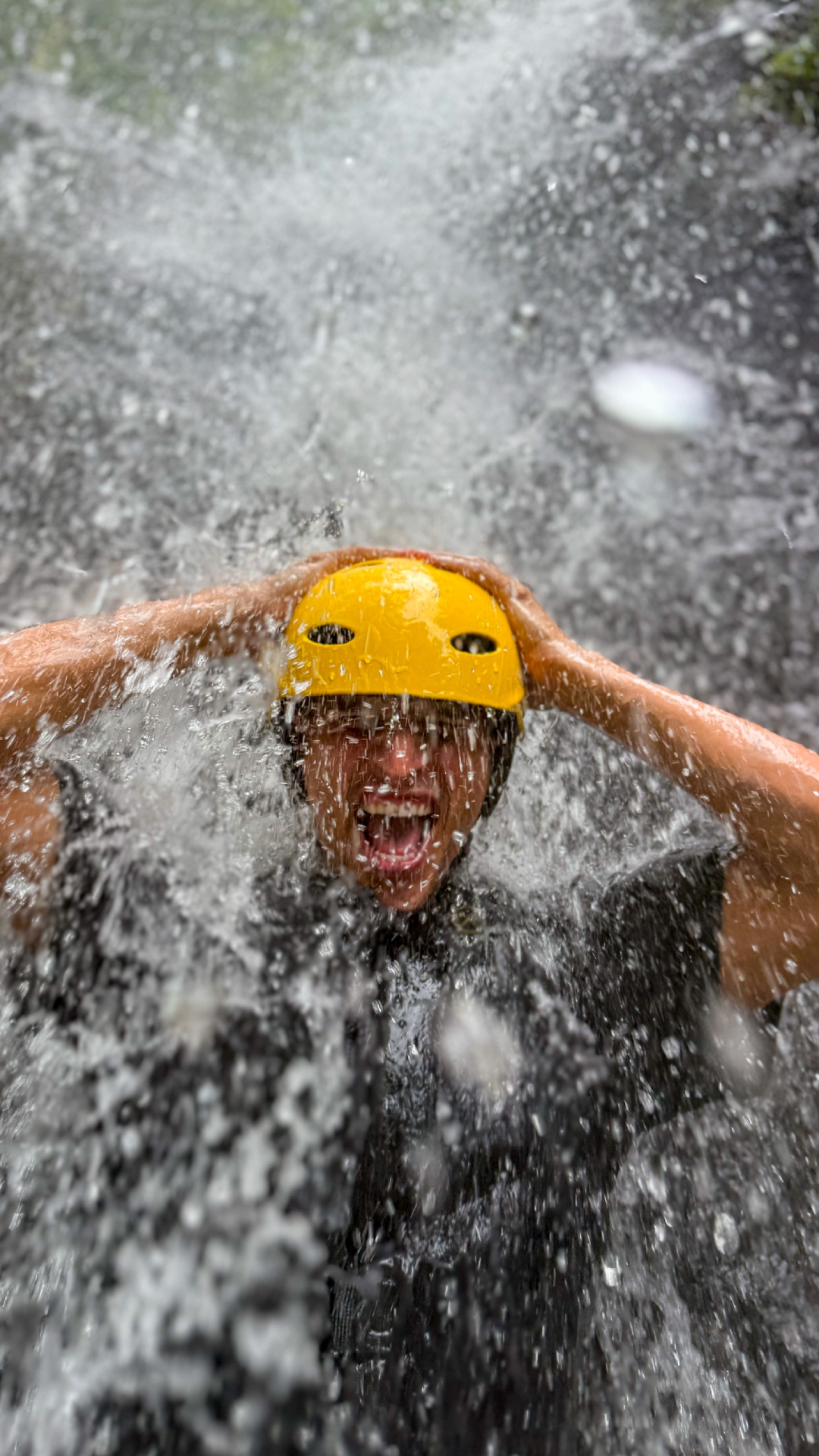 Person wearing a yellow helmet under a waterfall, with water splashing dramatically.
