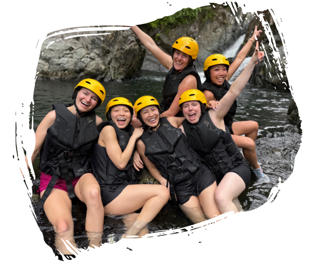 Group in life jackets and helmets, smiling by a rocky river.