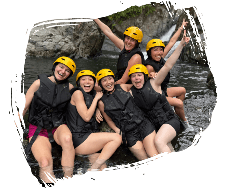 Group in life jackets and helmets, smiling by a rocky river.