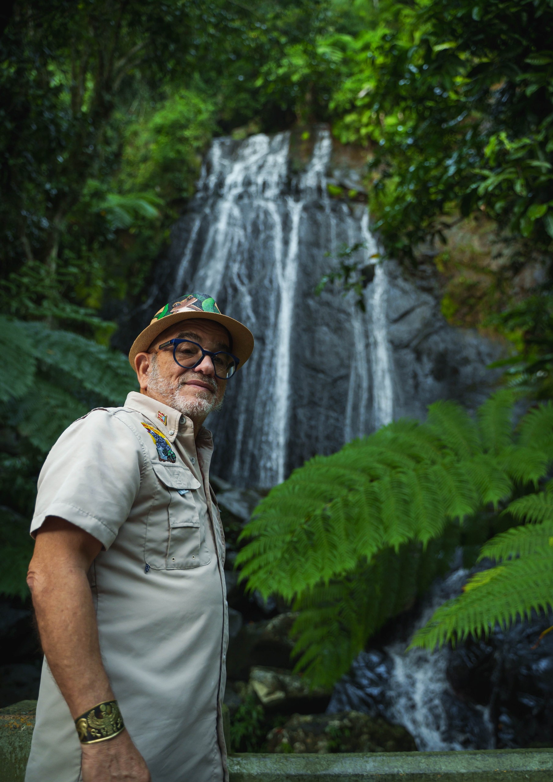 Man in hat stands in front of a waterfall in a lush forest.