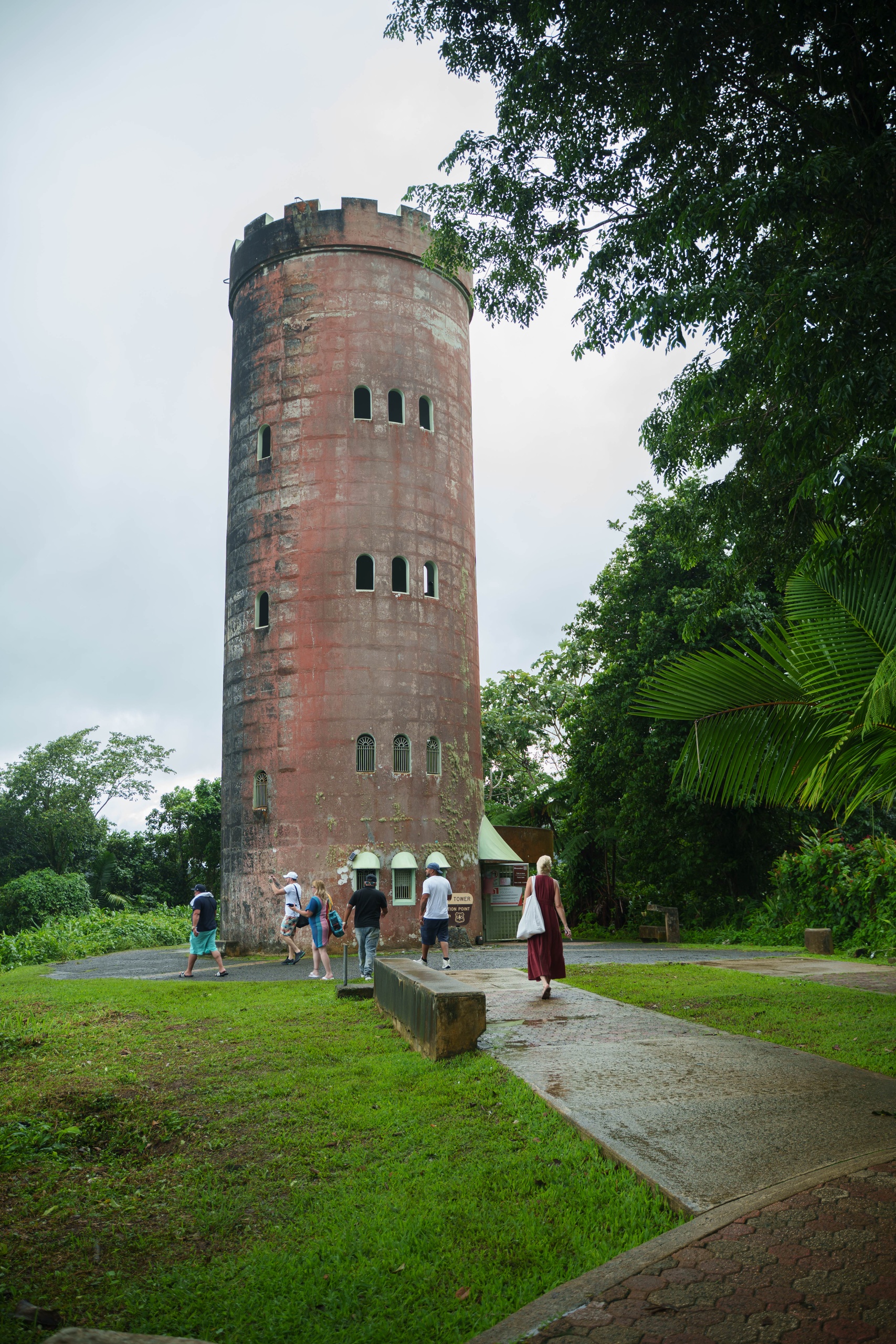 People visiting a tall, round, brick tower with small windows and lush greenery.