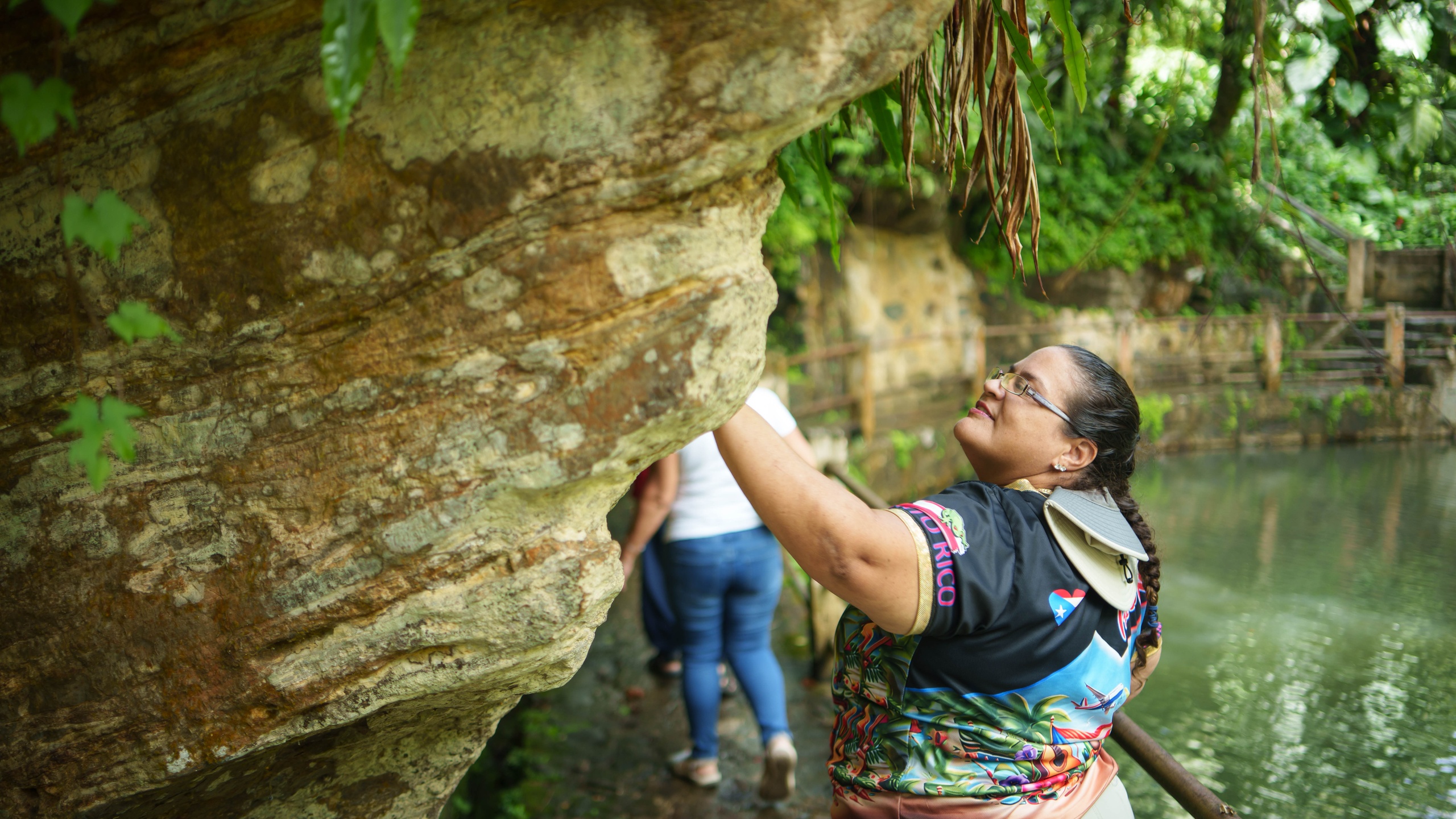 Woman smiling and touching a large rock in a lush, green outdoor setting.