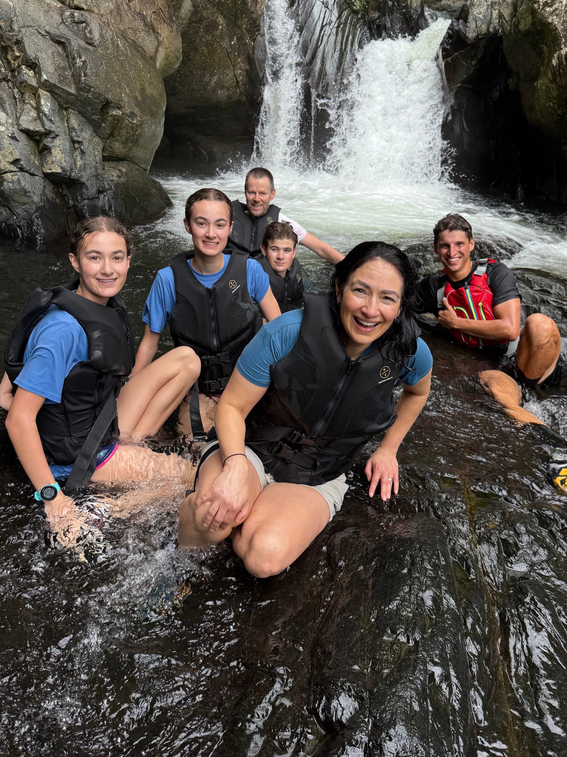 Group of six in life vests sit near a waterfall on rocks, smiling.