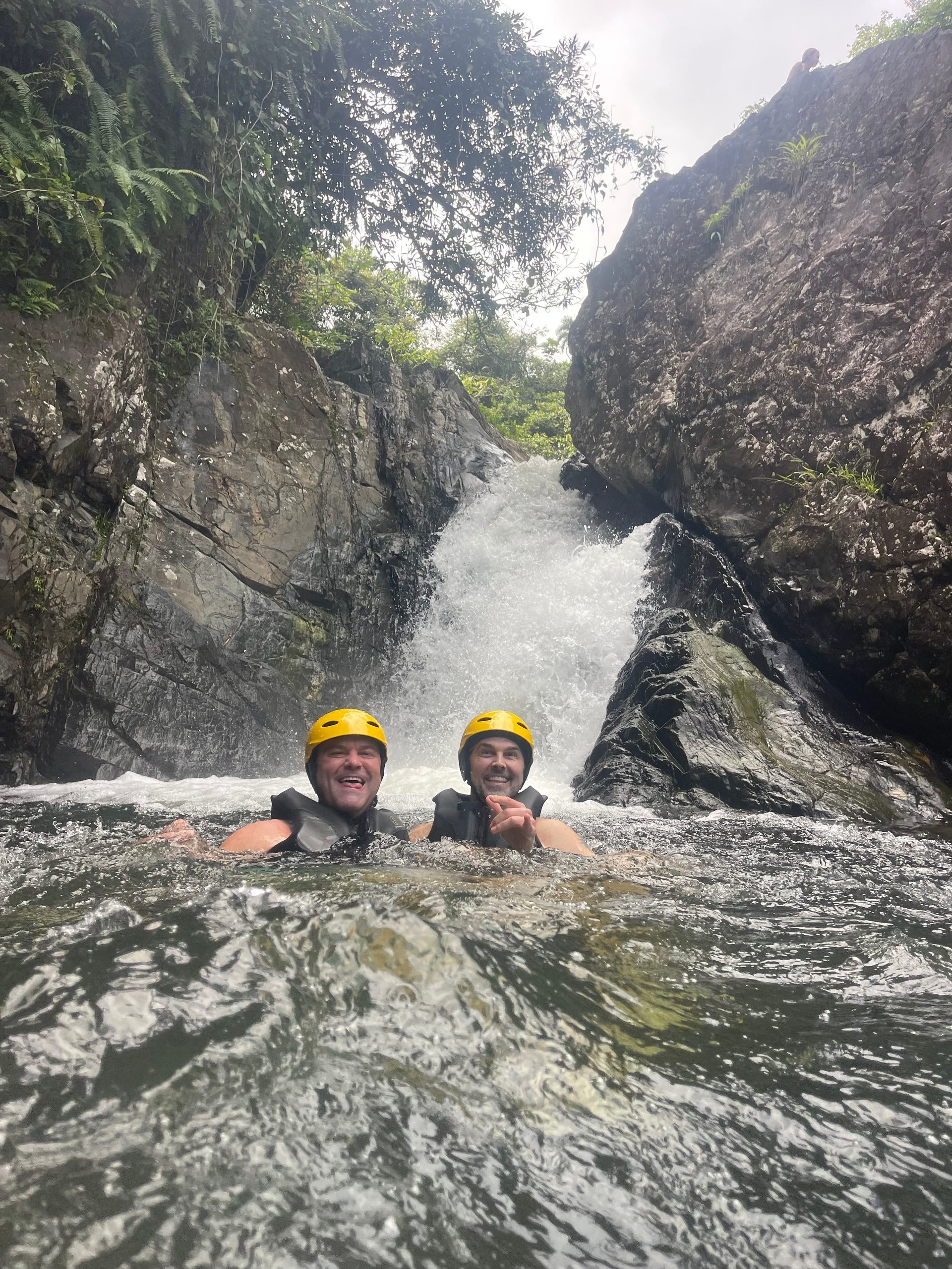 Two people with helmets smiling in a rocky river near a waterfall.