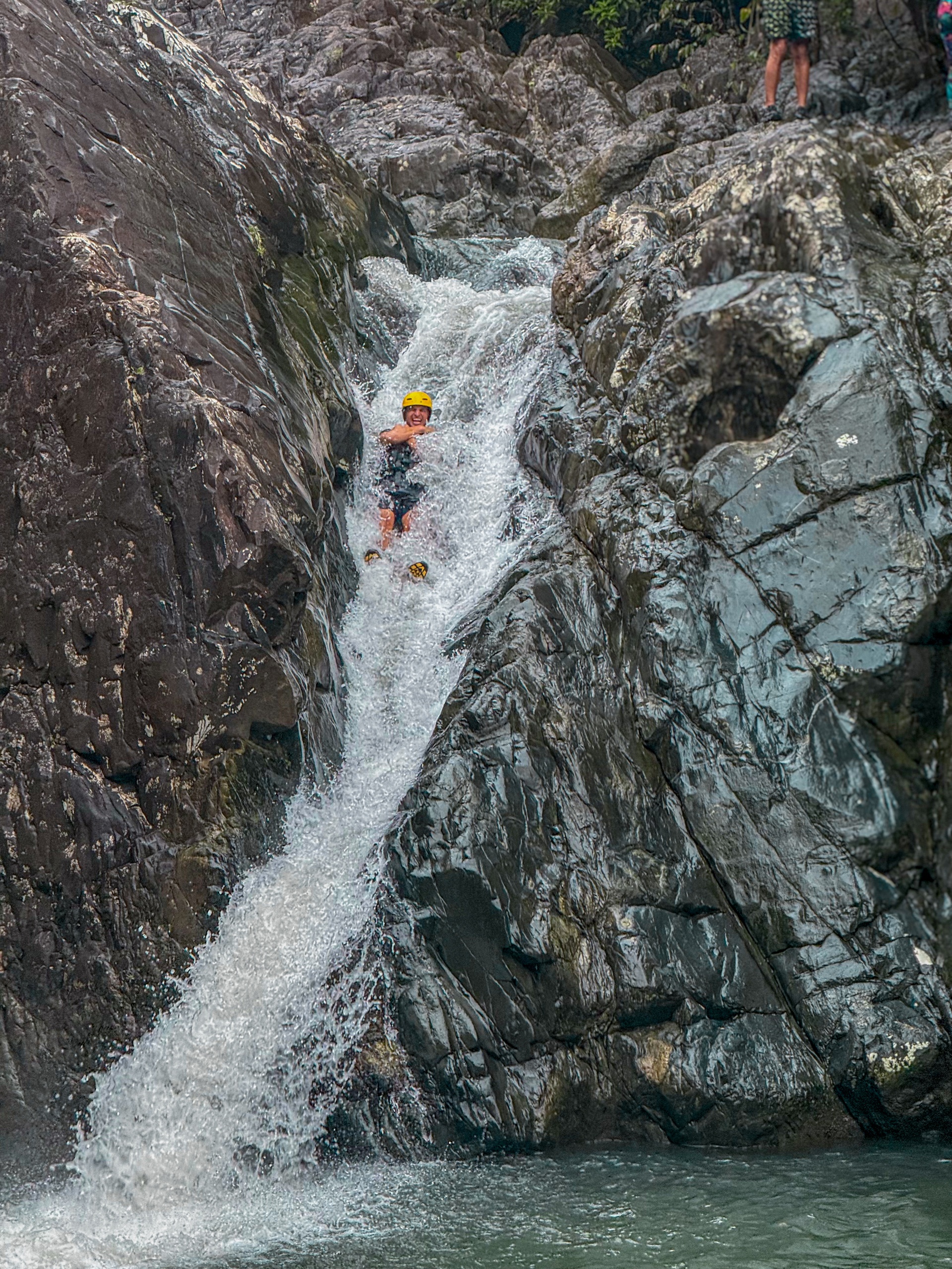 Person sliding down a rocky waterfall into a pool below.