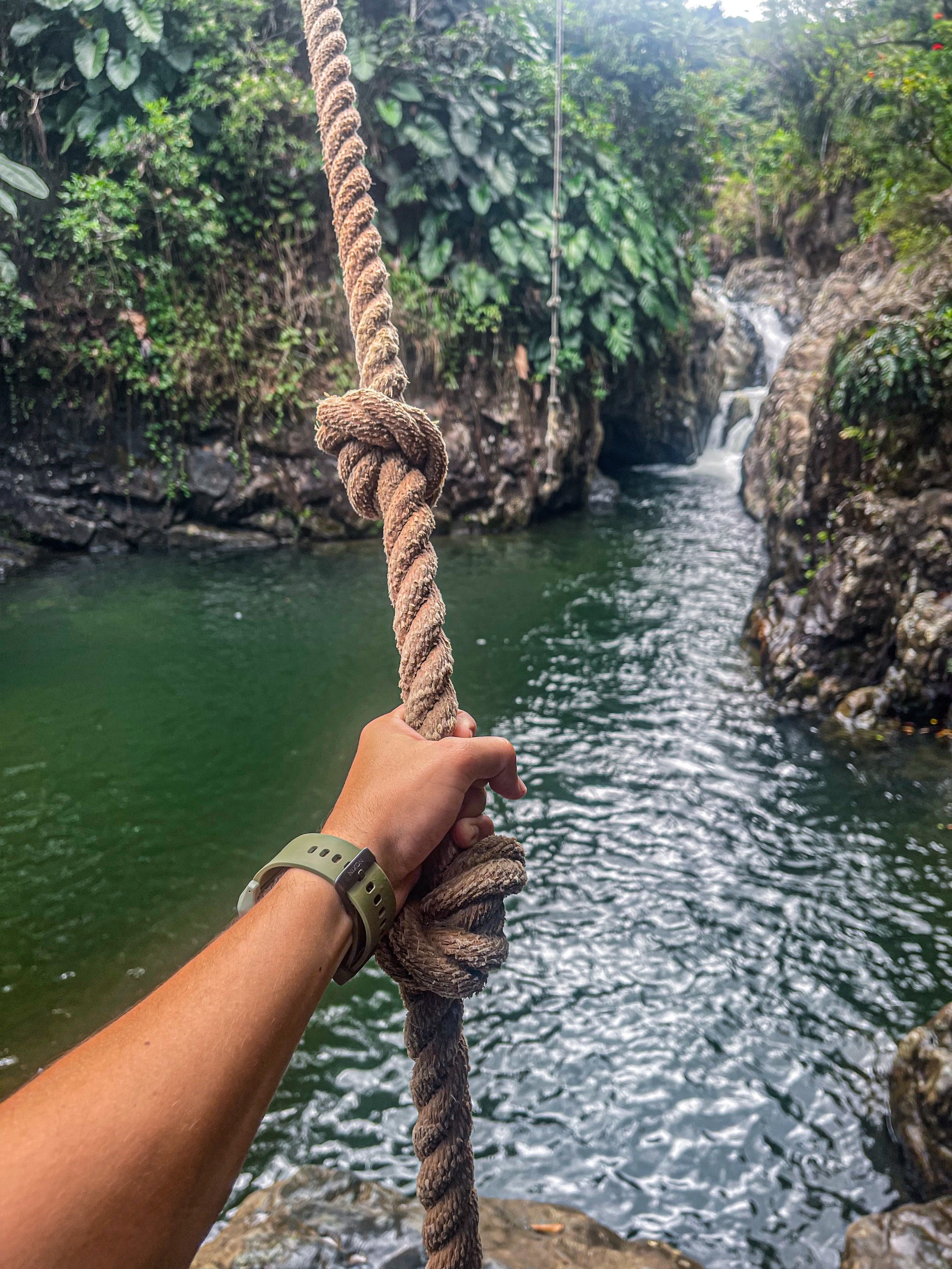 Hand holding rope over river with waterfall in lush, rocky landscape.