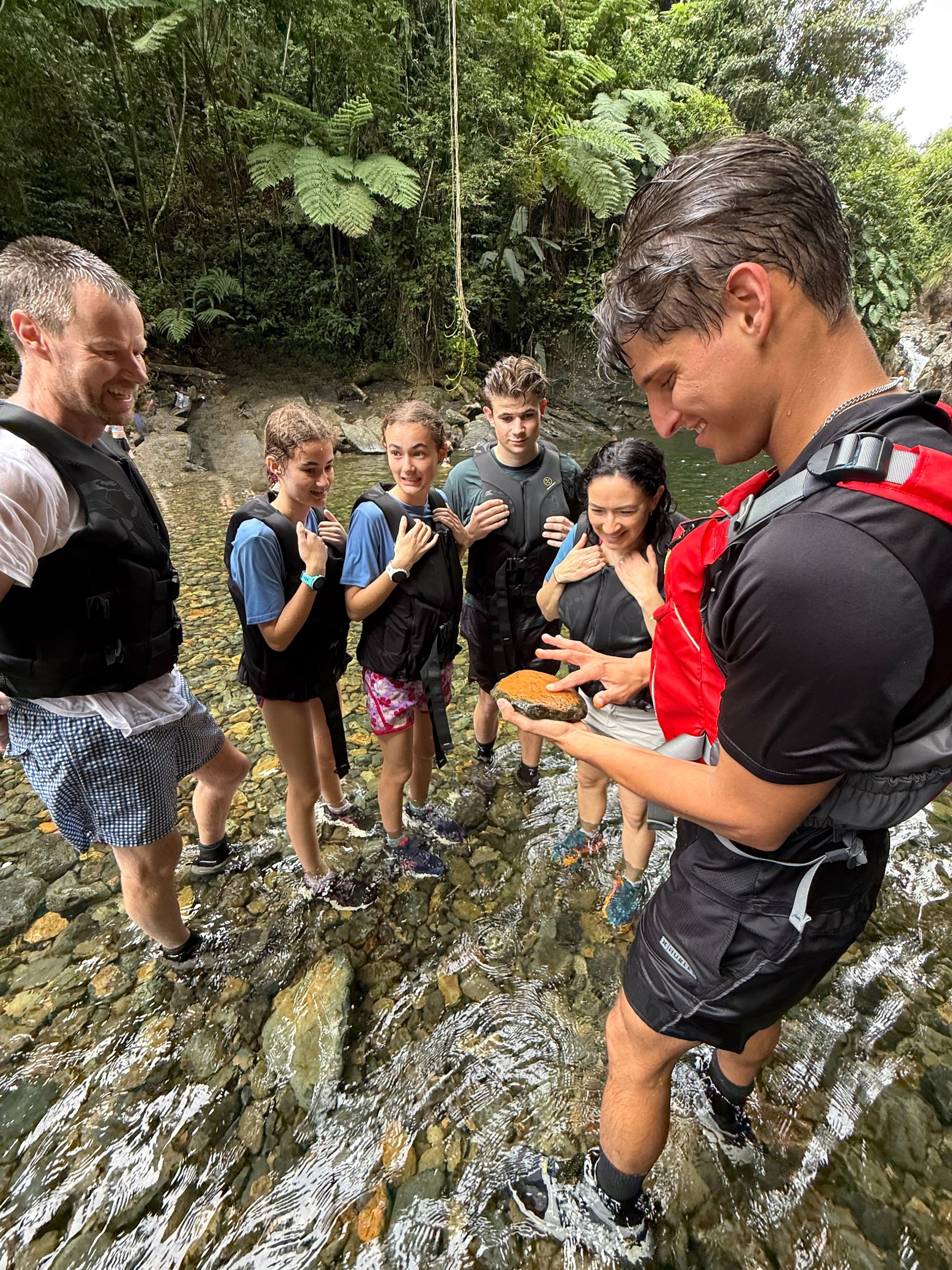 Group in shallow stream observes man holding object, surrounded by lush greenery.