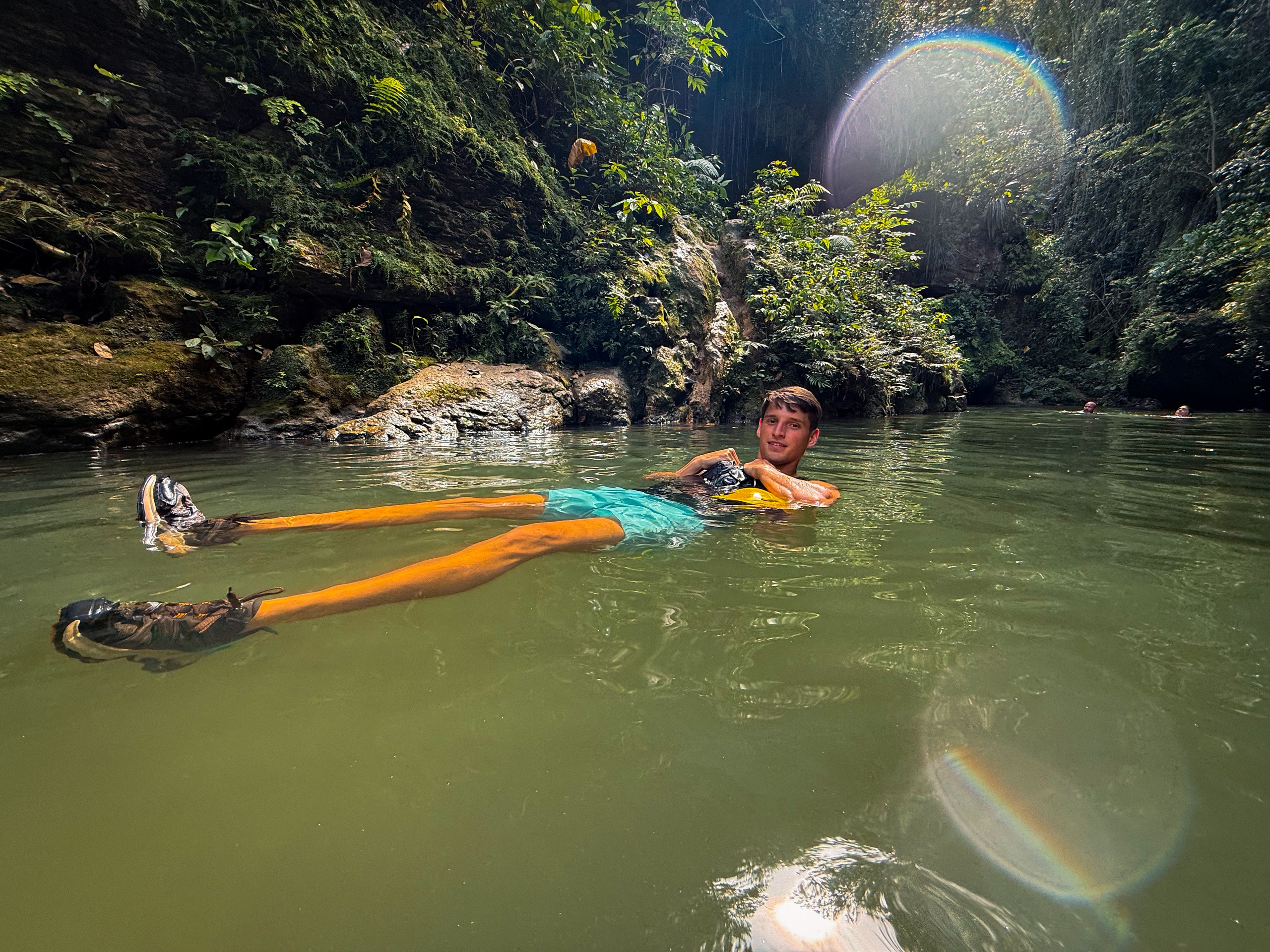 Person relaxing in a green river surrounded by lush vegetation and a lens flare rainbow.