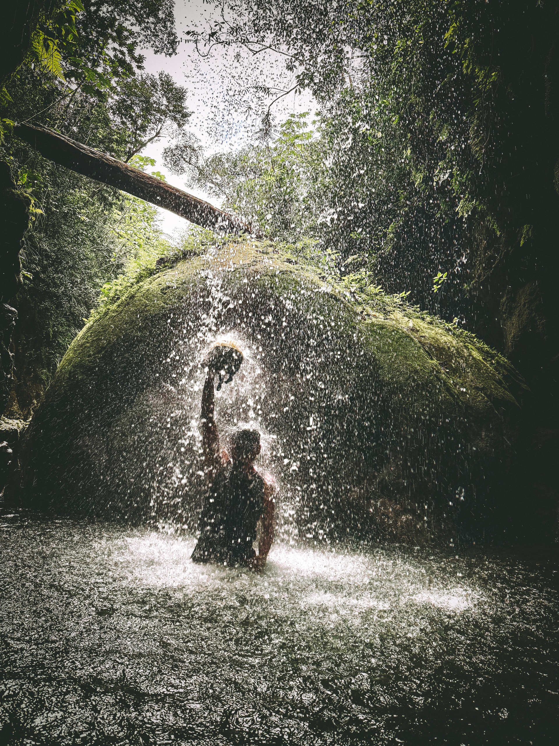 Person standing under a waterfall with arm raised, surrounded by lush greenery.