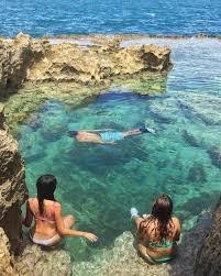 Two people sitting by a natural rocky pool with a swimmer in clear blue water.