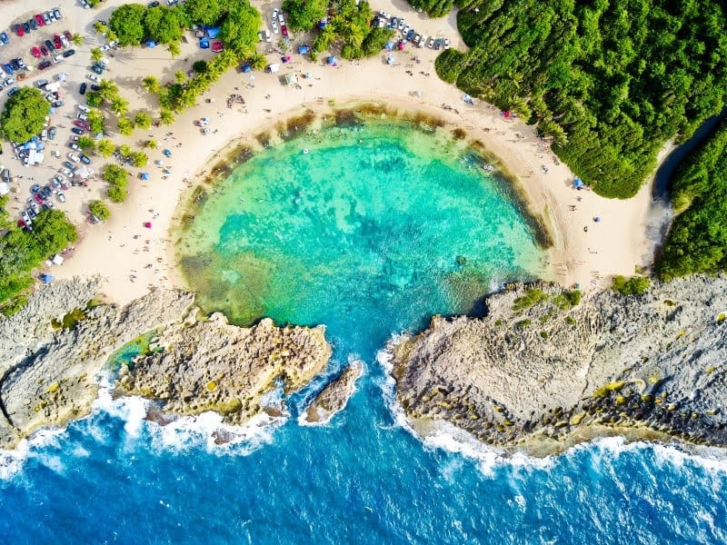 Aerial view of a circular beach cove with turquoise water and rocky coastline.