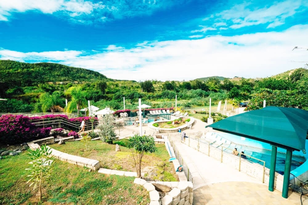 Scenic landscape with hills, pool, and surrounding greenery under a bright blue sky.