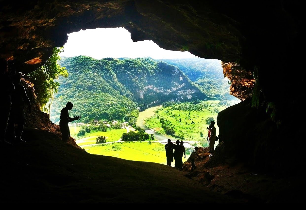 View from a cave opening overlooking a lush valley with people silhouetted in the foreground.