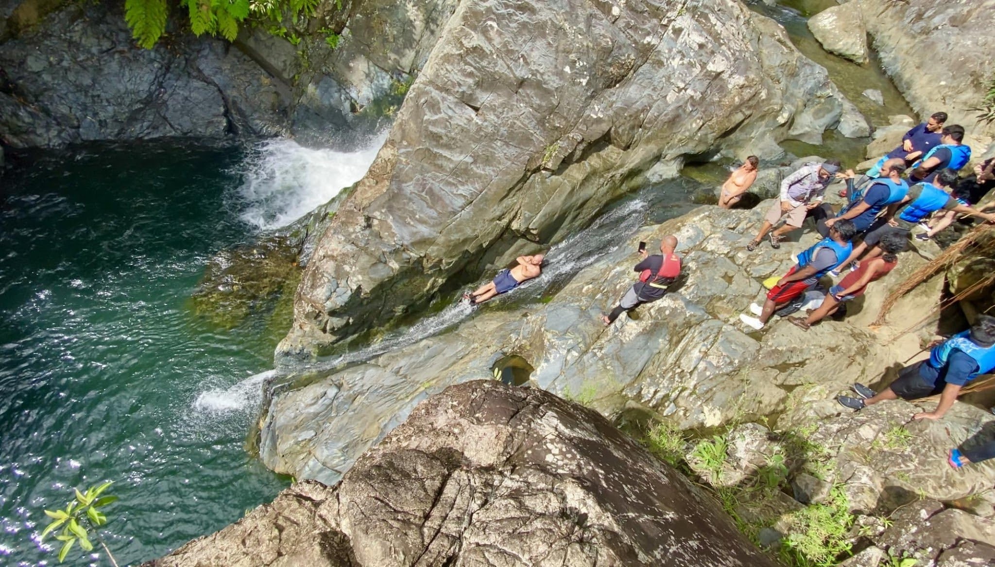 People in life vests sit on rocky slope by a pool of water near a waterfall.