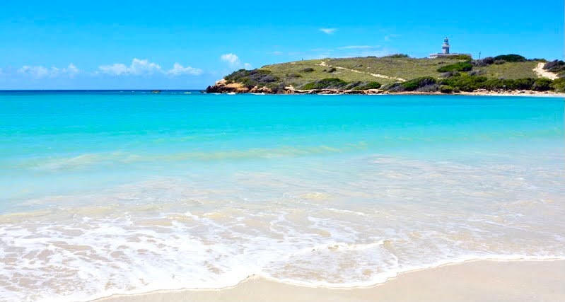 Sunny beach with turquoise water and a distant hill with a lighthouse.
