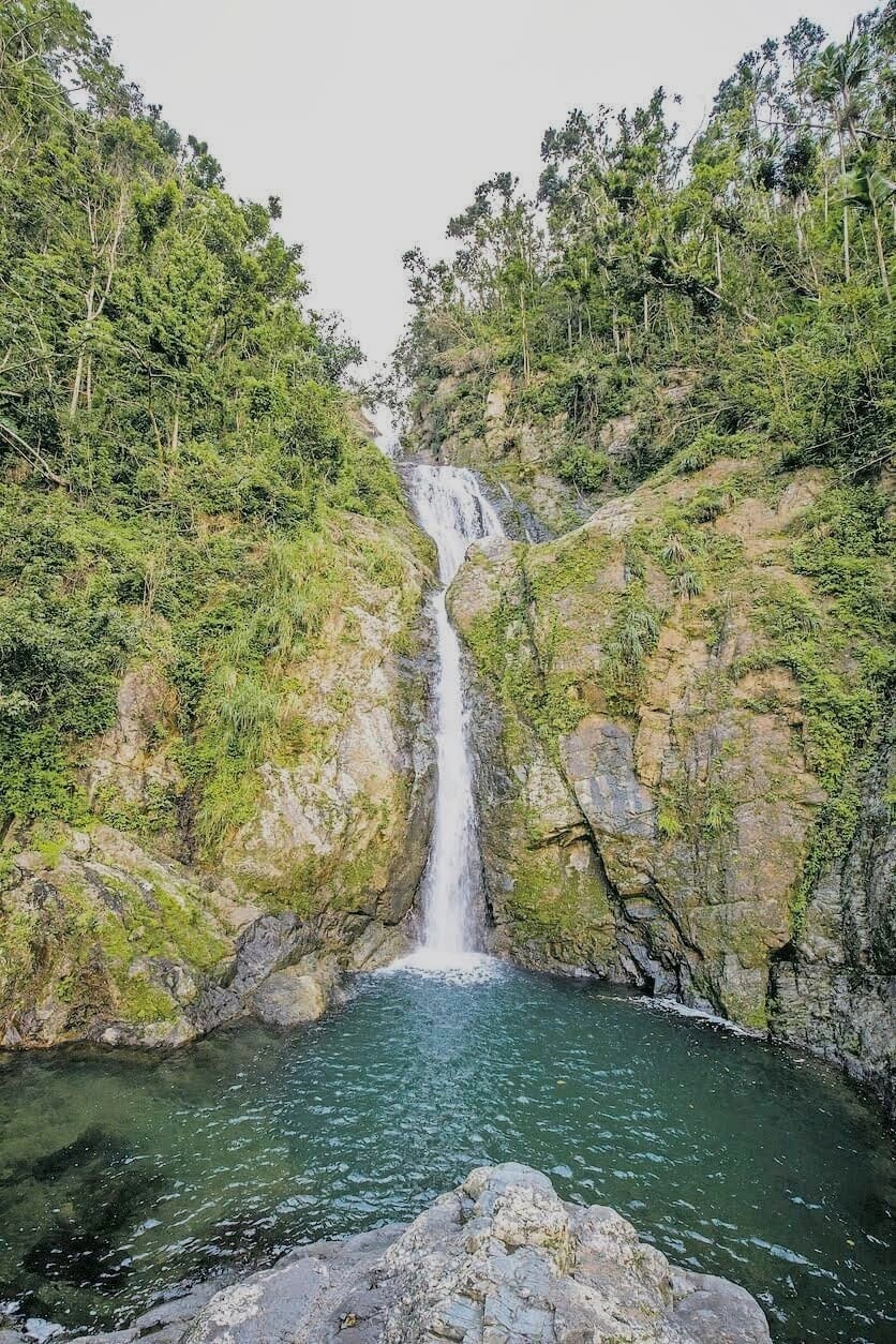 Tall waterfall cascading into a rocky pool surrounded by lush greenery.