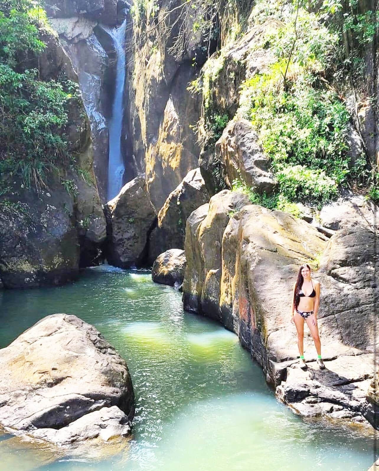 Woman in a bikini stands by a rocky waterfall pool.