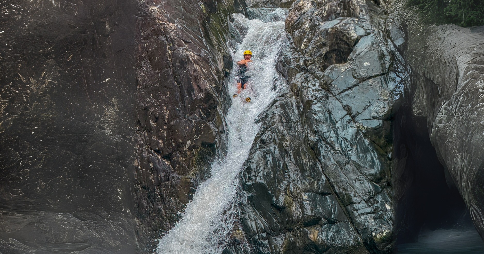Person wearing helmet slides down a narrow rocky waterfall.