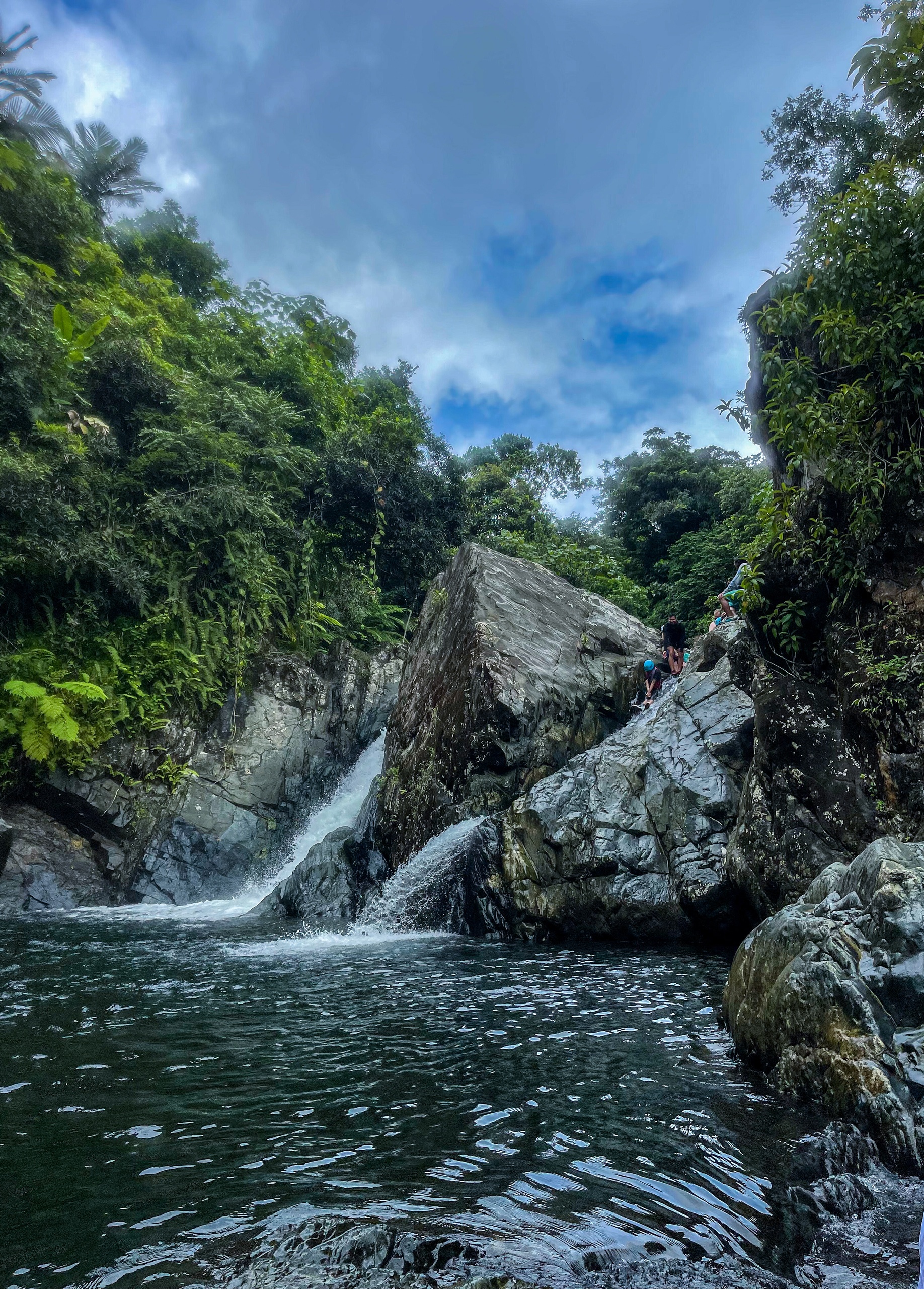 Waterfall cascading between rocks and lush green forest under a cloudy sky.