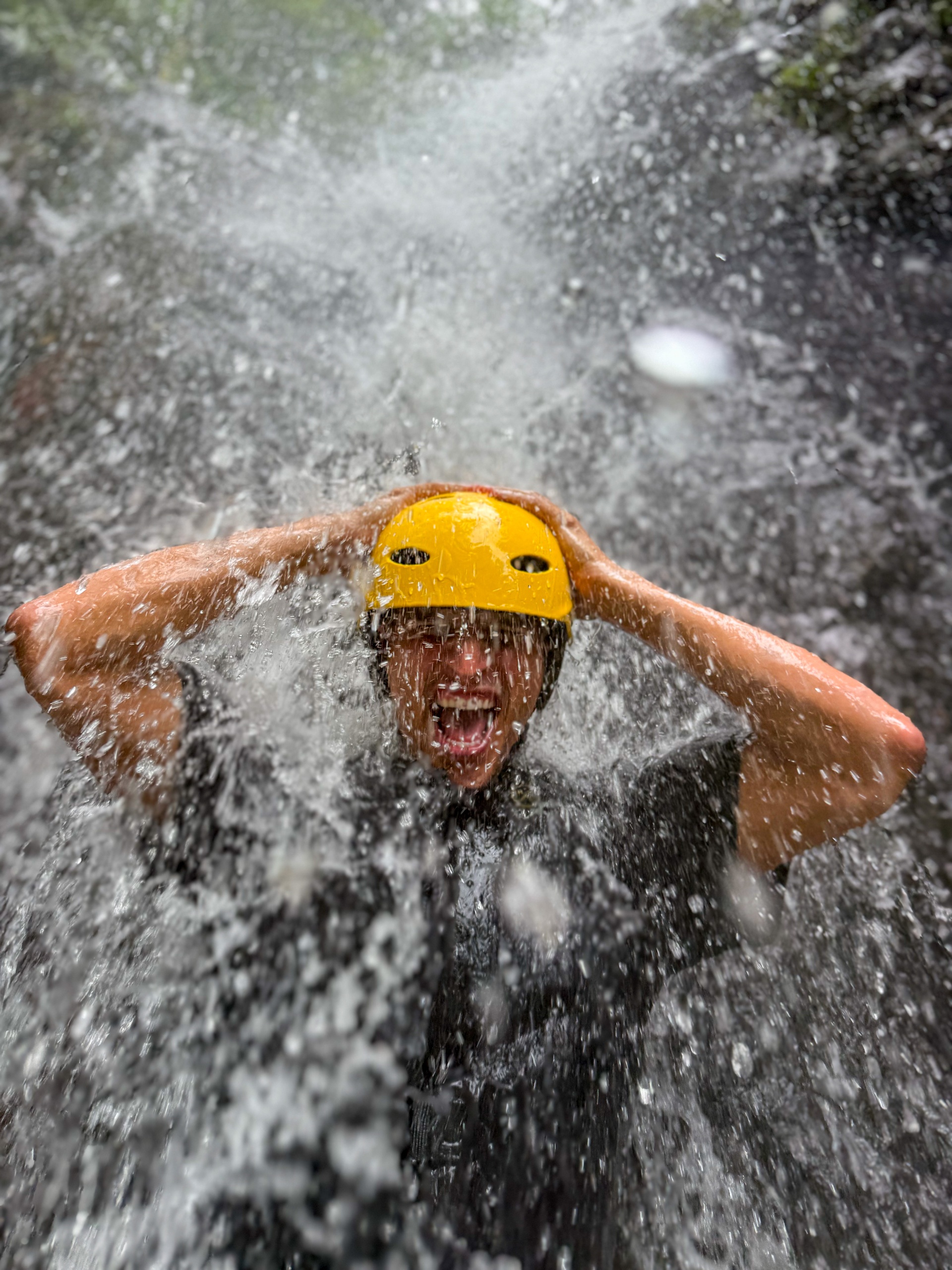 Person wearing a yellow helmet under a waterfall, hands on head, mouth open with water splashing around.