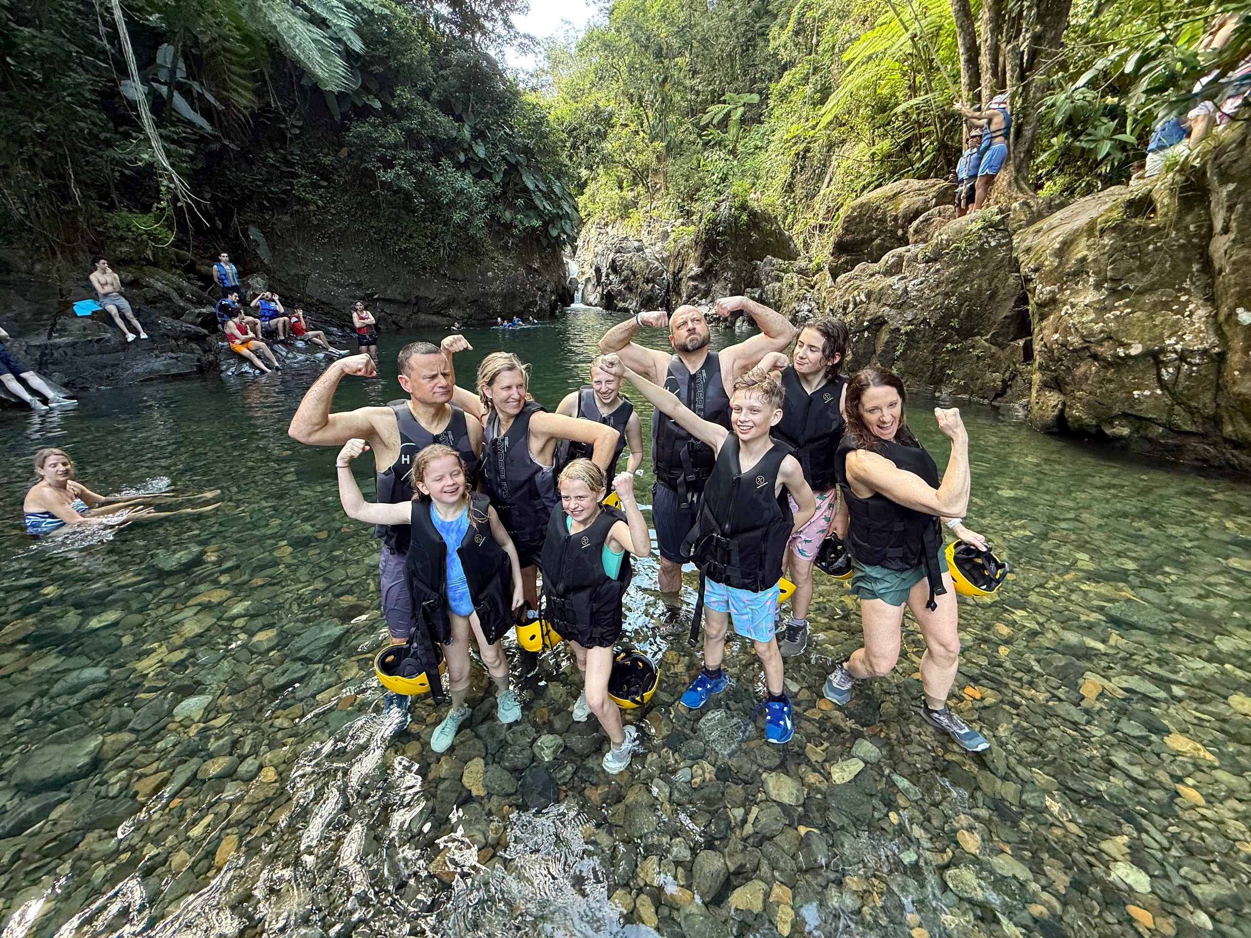 Group in life vests posing in a rocky river, flexing muscles, surrounded by trees.