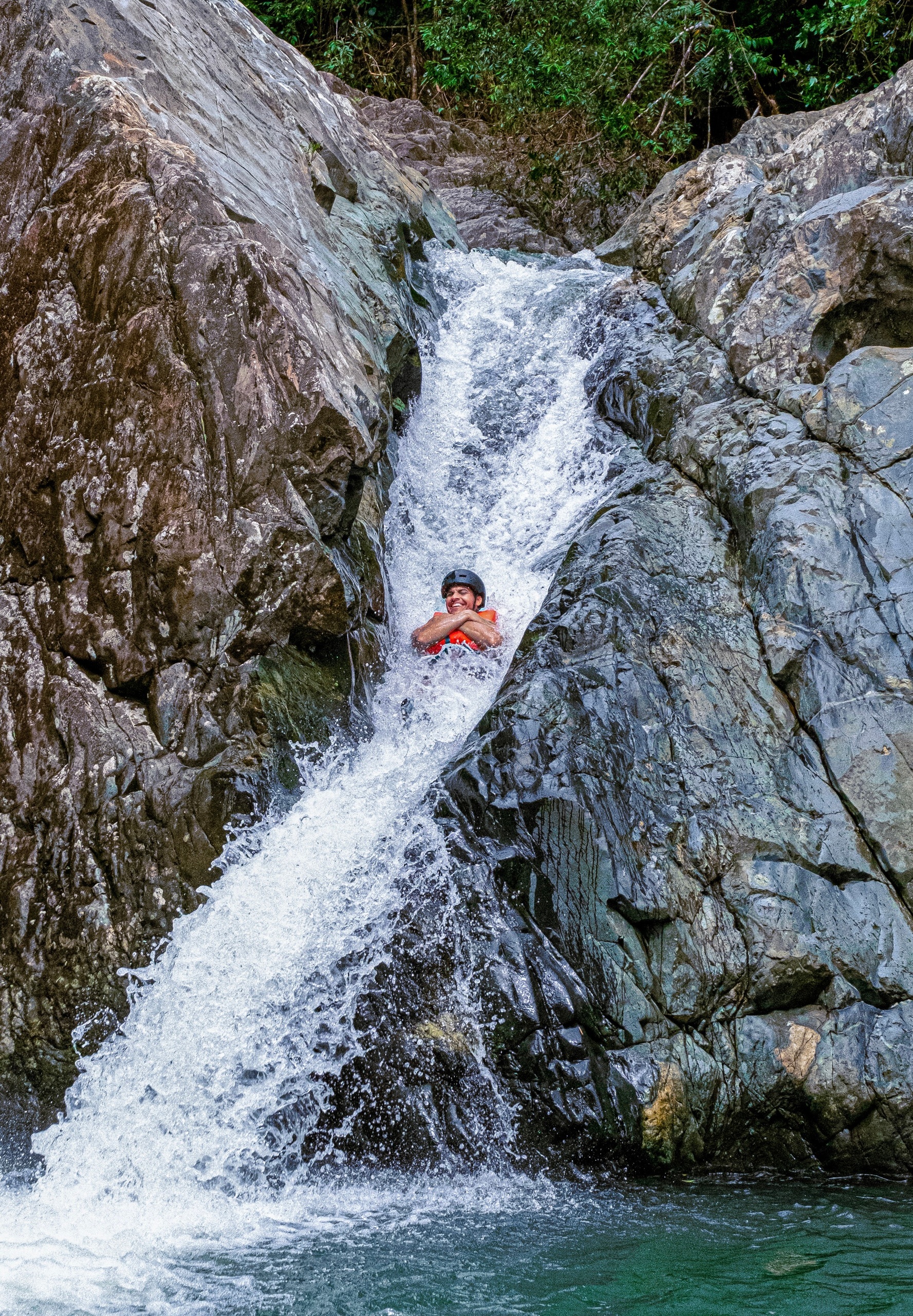 Person sliding down a rocky waterfall with lush greenery around.