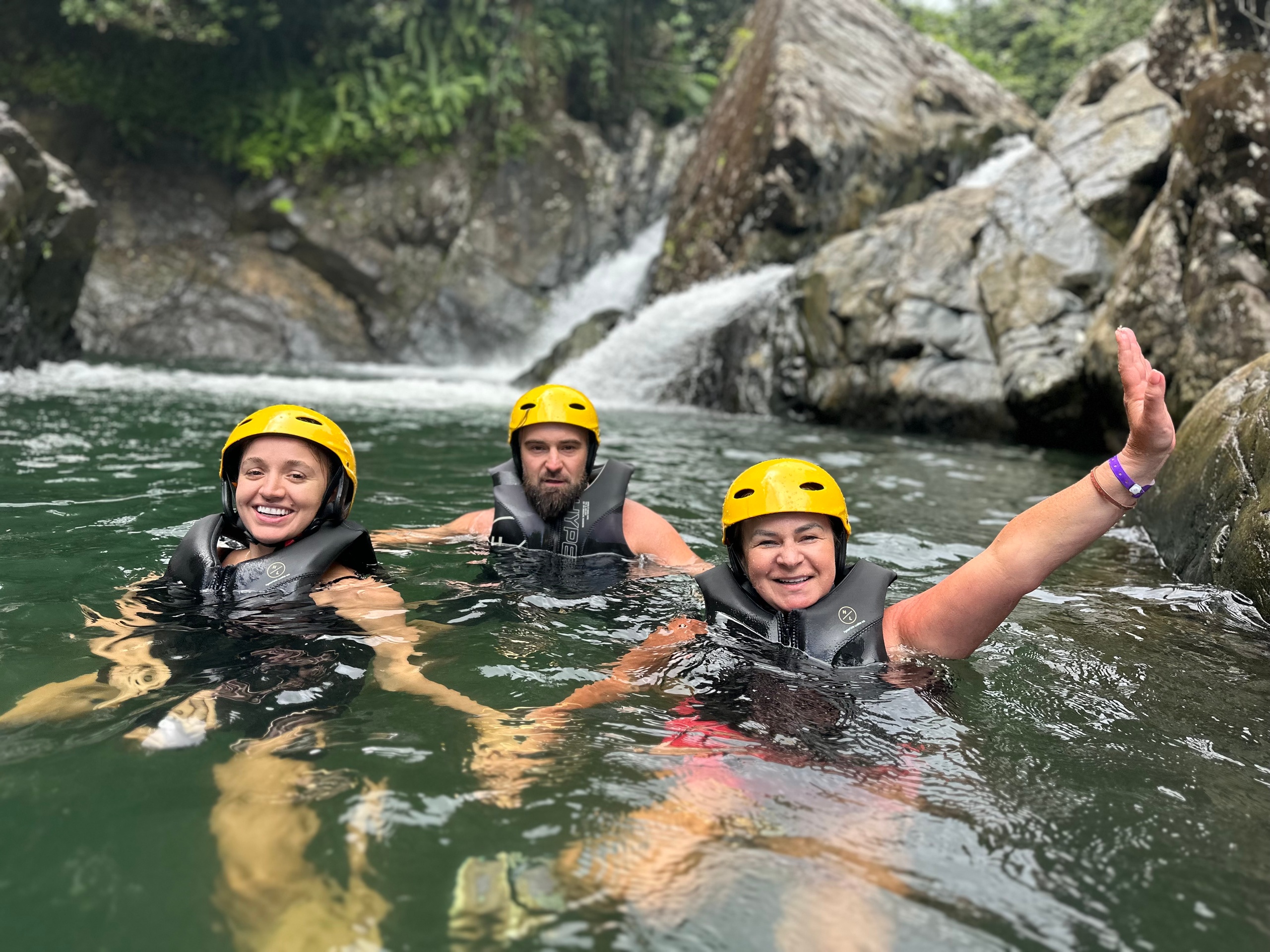 Three people in helmets and life jackets swimming near rocks and waterfall.