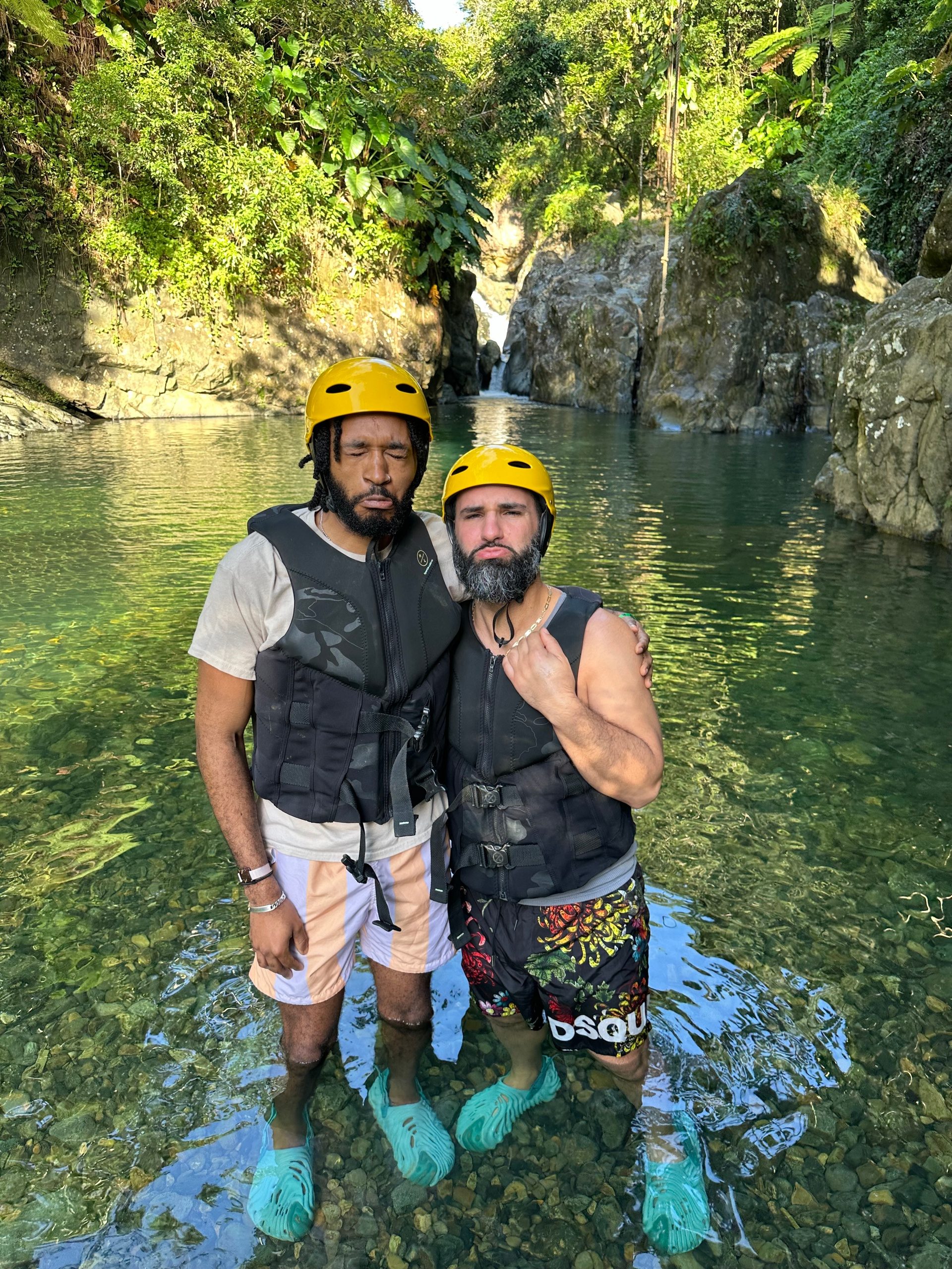 Two people in helmets and vests stand in a rocky stream with lush greenery.