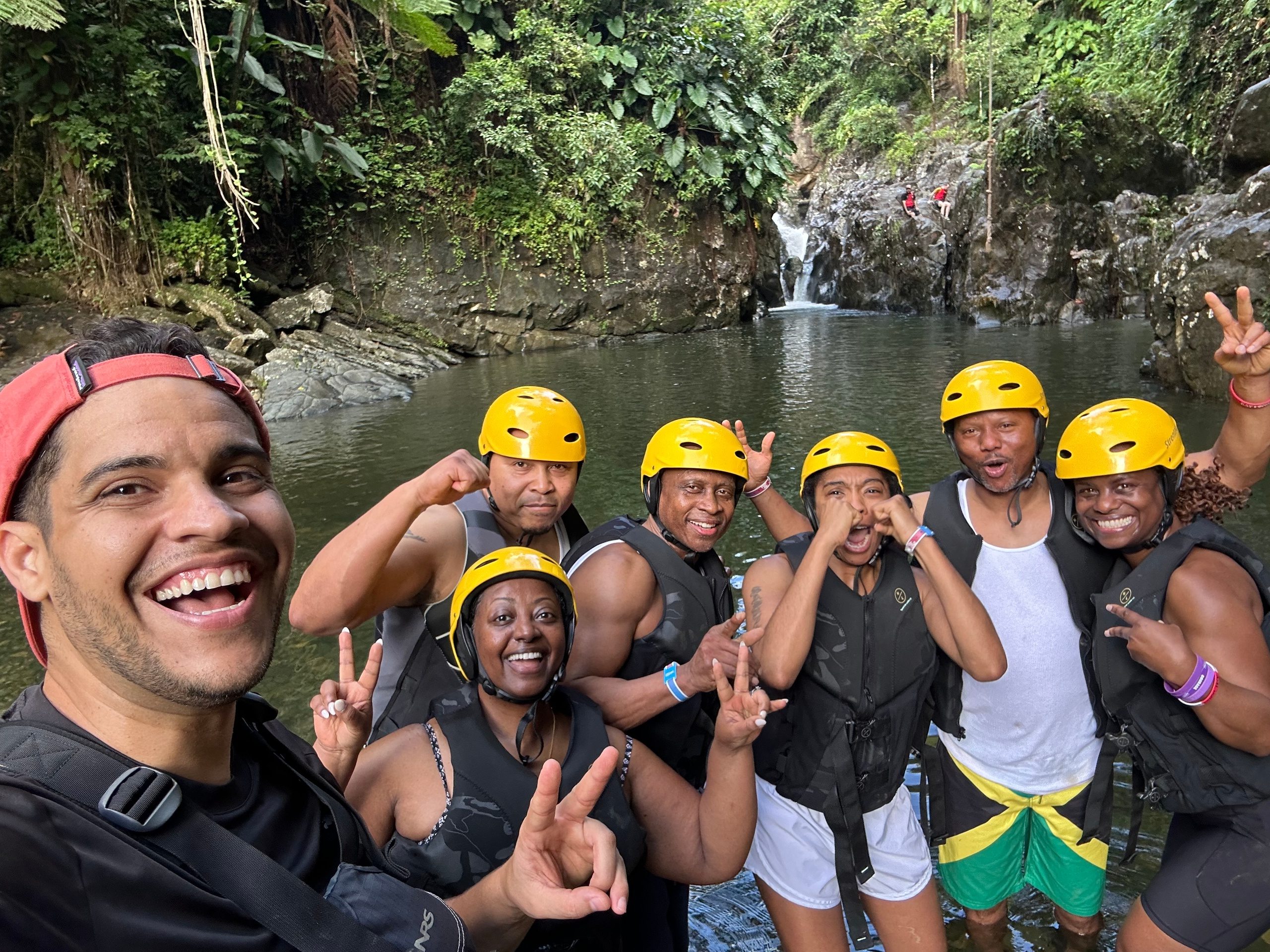 Group in helmets and vests posing by a river and waterfall, surrounded by greenery.